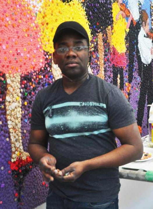 thumbnail-13.jpg Man standing in front of a colorful mosaic wall, holding art supplies.